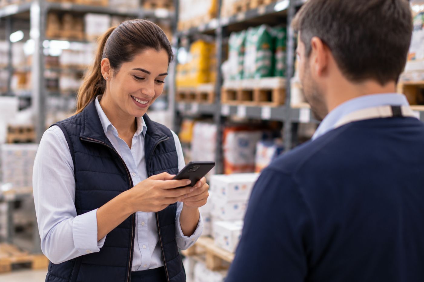 Vendedora registrando una visita en el celular mientras conversa con un cliente en un depósito